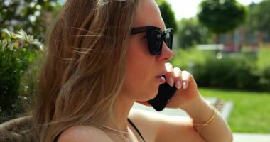 Kadın telefonu kulağına dayayıp dikkatlice dinlerken dudakları hafifçe yarılmış ve endişe duyuyor. Kadın şehir parkında güneşli bir günde ciddi telefon konuşmalarına odaklanıyor