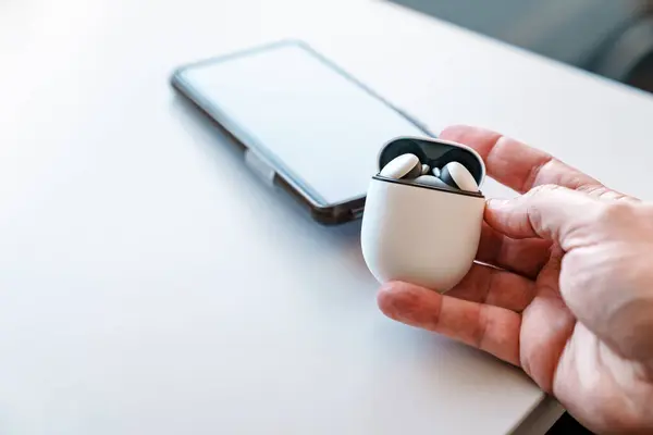 Wireless earbud headphones in their charging case with the lid open, being held by a caucasian adult male hand. A smartphone is in the background sitting on a white desk. Room for copy space.