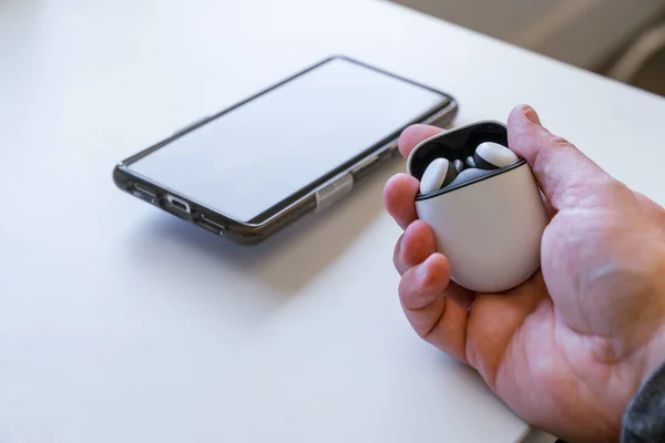 Wireless earbud headphones in a charging case being held by a caucasian male hand. A smartphone is in the background on the desk.