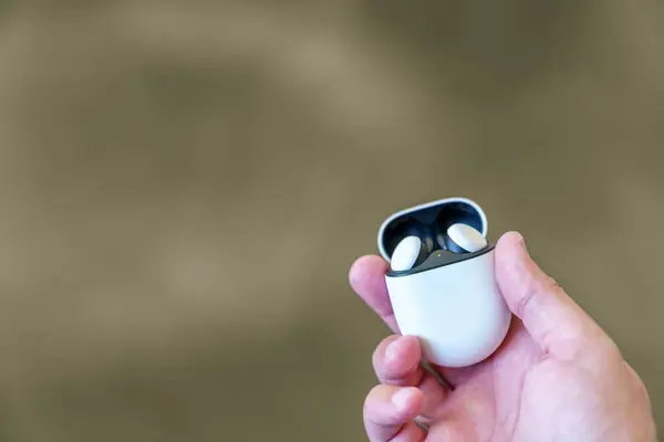 Wireless earbuds headphones in their charging case with the lid open, being held by a caucasian adult male hand. Blurred brown background and room for copy space.