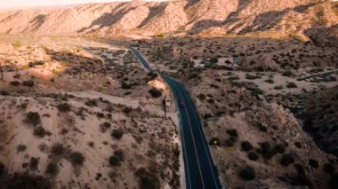 Pioneertown, California yolunda giden tek bir kırmızı aracın hava aracı görüntüsü. Güneş arka plandaki tepelerden batıyor..