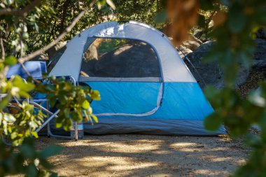 Ormanlık bir alanda mavi ve gri bir çadır var. Çadırın önünde iki sandalye ve sandalyelerden birinde bir su şişesi var..