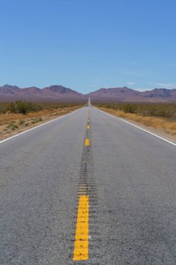 Sarı çizgileri olan uzun, düz bir asfalt yol çöldeki açık mavi gökyüzünün altındaki uzak, alçak dağlara doğru uzanır..