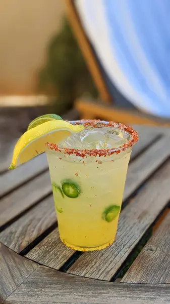 Buzlu, limonlu, jalapenolu ve jalapenolu şeffaf plastik bir bardakta duran bir Mango Jalapeno margarita içeceği, açık havada eskimiş ahşap bir masada oturur..