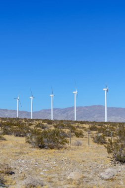 Açık mavi gökyüzü ve kurak Palm Springs çöl manzarasına karşı beş beyaz rüzgar türbini bir sırada duruyor. Arka planda görünen dağlarda kopyalama alanı.