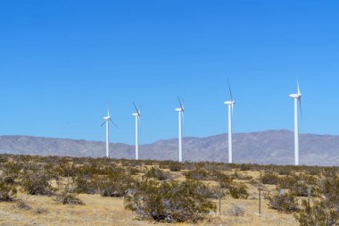 Beş beyaz rüzgar türbini, Palm Springs 'in kurak çöl manzarasında ve açık mavi gökyüzünün altındaki uzak dağlarda bir sırada duruyor. Boşluğu Kopyala.