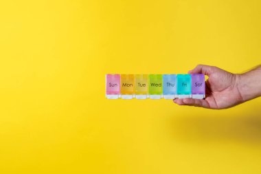 Gökkuşağı renklerinde, erkek eli sarı arka planda ve fotokopi alanı olan haftalık 7 günlük hap organizatörü