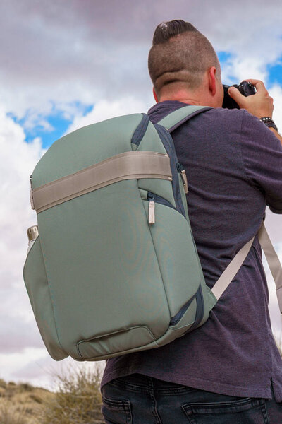 Зеленый рюкзак на спине во время съемки камерой в пустыне с голубыми облаками