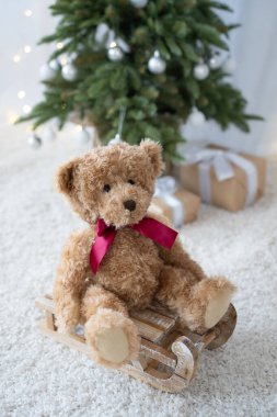 Kahverengi renkli oyuncak ayı, arka planda Noel ağacı ve Noel ışıkları olan ahşap kızağın üzerinde oturuyor..