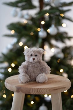 Bir Noel ağacının yanındaki tahta taburede oturan açık kahverengi bir oyuncak ayı. Noel teması.