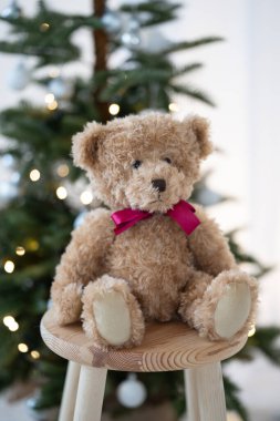 Kırmızı kurdeleli açık kahverengi bir oyuncak ayı Noel ağacının yanındaki tahta taburede oturuyor. Noel teması.