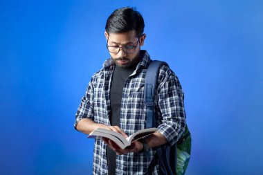 Bir üniversite öğrencisi kitabı okuyor ve mavi arka planda izole edilmiş sırt çantası taşıyor..