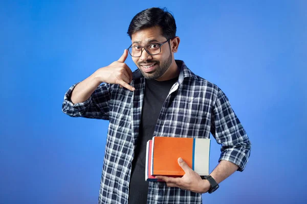 Portrait of a college male student showing call me hand gesture with cheerful smile, isolated on blue background.