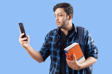 Genç Hintli erkek kolaj öğrencisi cep telefonu ve tek renk mavi arka plana karşı duran kitaplar kullanıyor.