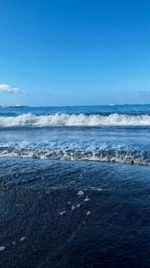 Atlantic waves hit the sandy beach. Black sand and blue sky. Vertical image. Costa Adeje, Spain. High quality photo clipart