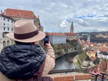 Şapkalı bir bayan turist, Cesky Krumlov 'un cep telefonuyla fotoğraflarını çekiyor. Eski bir peri masalı kasabası. Büyülü atmosfer ve muhteşem gökyüzü. Yüksek kalite fotoğraf. 