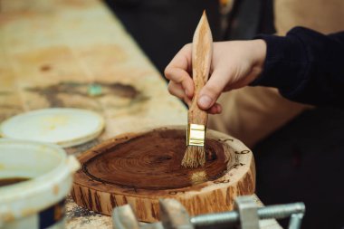 Boy is putting a protective mordant on the wooden disk, closeup. Young carpenter working with wood in craft workshop. Boy makes wooden clock in the workshop.