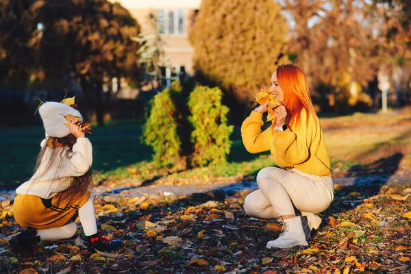 Family enjoying the autumn vacation. Stylish family on autumn walk. Happy mother and child throwing autumn leaves in the park. Mom and daughter having fun together. Autumn fashion, lifestyle and vacation.