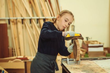Young carpenter working with wood and sandpaper in craft workshop. School, development and learning concept. Cute boy makes wooden clock in the workshop.