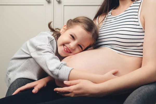 Happy little girl touching her pregnant mom tummy. Pregnant mother and daughter spending time together at home. Sweet daughter hugging mommy tummy. Happy family, expecting baby birth.