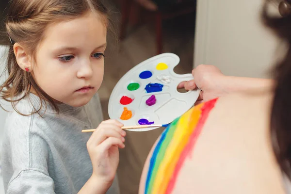 Happy little girl drawing rainbow on pregnant belly her mother. Baby birth expecting time and belly painting. Pregnant mom and child having fun together at home.