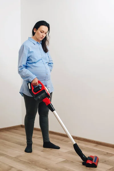 Pregnant woman cleaning floor with handheld vacuum cleaner. Young pregnant woman enjoys cleaning her house. Easy cleaning with a wireless vacuum cleaner. Modern easy cleaning.