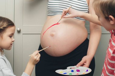 Baby birth expecting time and belly painting. Happy children and pregnant mom having fun together at home. Family, healthy pregnancy and baby birth. Happy kids painting pregnant belly their mother.