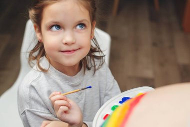 Sweet little daugter painting pregnant belly their mother. Baby birth expecting time and belly painting. Pregnant mom and her child having fun together at home. Family, healthy pregnancy and baby birth.