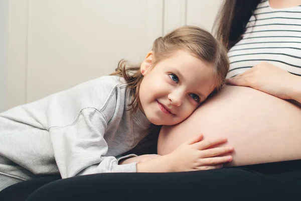 Happy little girl touching her pregnant mom tummy. Pregnant mother and daughter spending time together at home. Sweet daughter hugging mommy tummy. Happy family, expecting baby birth.