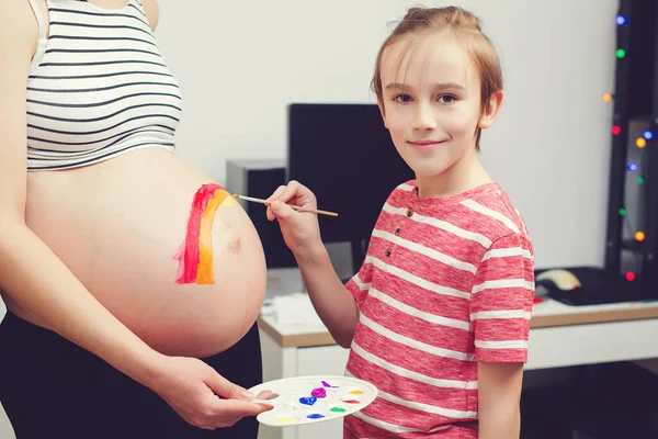 Cute boy drawing rainbow on pregnant belly his mother. Baby birth expecting time and belly painting. Pregnant mom and her son having fun together at home. Family, healthy pregnancy.