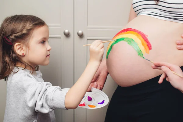 Sweet little daugter painting pregnant belly their mother. Baby birth expecting time and belly painting. Pregnant mom and her child having fun together at home. Family, healthy pregnancy and baby birth.