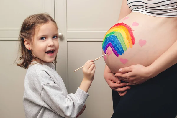 Sweet little daugter painting pregnant belly her mother. Baby birth expecting time and belly painting. Pregnant mom and her child having fun together at home. Family, healthy pregnancy and baby birth.