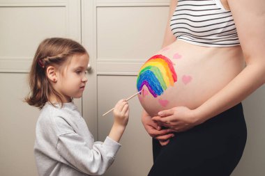 Sweet little daugter painting pregnant belly her mother. Baby birth expecting time and belly painting. Pregnant mom and her child having fun together at home. Family, healthy pregnancy and baby birth.