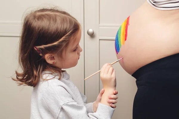 Sweet little daugter painting pregnant belly her mother. Baby birth expecting time and belly painting. Pregnant mom and her child having fun together at home. Family, healthy pregnancy and baby birth.