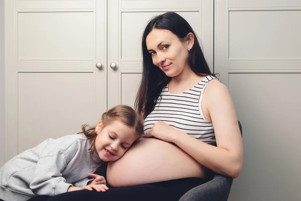 怀孕的母亲和女儿一起呆在家里 可爱的女儿抱着妈妈的肚子幸福的家庭 期待着孩子出生 快乐的小女孩摸她怀孕的妈妈肚子 — 图库照片