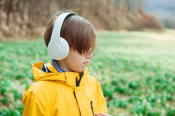 Sad boy listening to music at nature. Kid with headphones relaxing in the spring park. Upset boy with headphones on the walk. Lifestyle, childhood and emotions.
