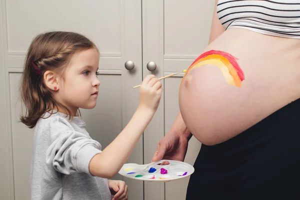 Sweet little daugter painting pregnant belly their mother. Baby birth expecting time and belly painting. Pregnant mom and her child having fun together at home. Family, healthy pregnancy, baby birth.