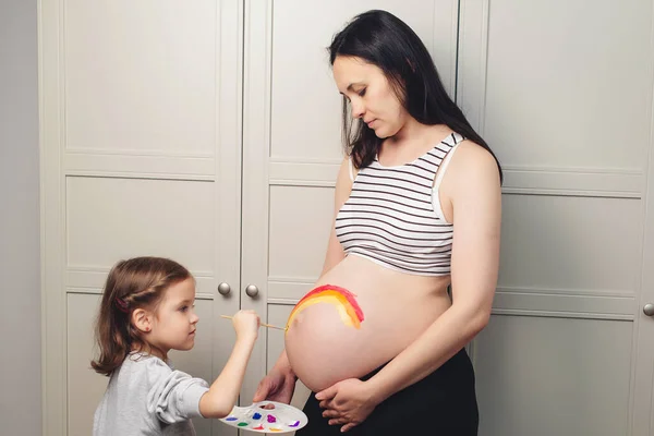 Sweet little daugter painting pregnant belly their mother. Baby birth expecting time and belly painting. Pregnant mom and her child having fun together at home. Family, healthy pregnancy, baby birth.