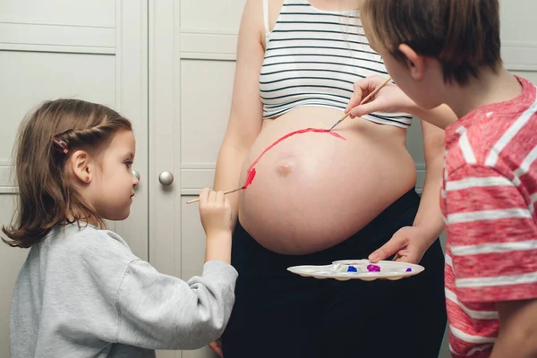 Happy kids painting pregnant belly their mother. Baby birth expecting time and belly painting. Happy children and pregnant mom having fun together at home. Family, healthy pregnancy and baby birth.