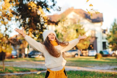 Mutlu çocuk doğa yürüyüşünde sonbahar yapraklarıyla oynuyor. Küçük kız kalın örülü kazak giyiyor. Sonbahar çocukları moda, yaşam tarzı.