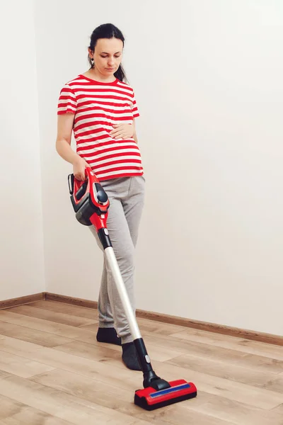 Young pregnant woman enjoys cleaning her house. Easy cleaning with a wireless vacuum cleaner. Pregnant woman cleaning floor with handheld vacuum cleaner. Modern easy cleaning.