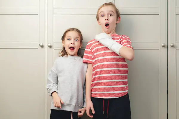 Boy broken arm, girl and boy is in shock. Shocked surprised boy with arm in sling. Boy has a cast on his arm. Child girl holding his brother\'s broken arm.