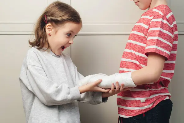 Boy broken arm, girl is in shock. Child girl holding his brother\'s broken arm. Boy holds hand bent broken arm cast on his arm.