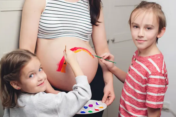 Baby birth expecting time and belly painting. Happy children and pregnant mom having fun together at home. Family, healthy pregnancy and baby birth. Happy kids painting pregnant belly their mother.