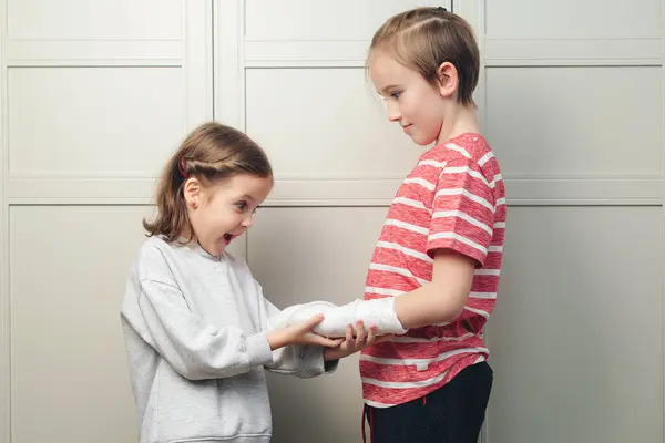Boy two broken arms, girl is in shock. Shocked surprised boy with arm in sling. Boy has a cast on his arm. Child girl holding his brother\'s broken arm. Boy holds hand bent broken arm cast on his arm.