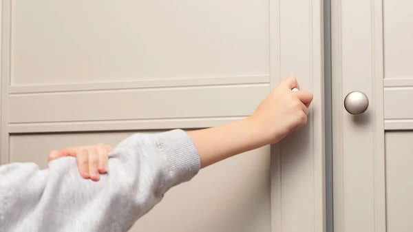 Child\'s hand opens the closet door. Cute baby girl playing with a wooden cupboard. Toddler baby opens the closet door in the home living room.