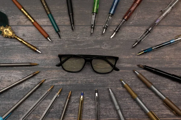 opticians and studio, intellectual forming an oval of pens, pens, markers surrounding a black glasses on a wooden table. with space for text.