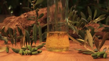 Zeytin yağı cam bir şişeye dökülür, kabarcıklar oluşur, etrafında yapraklar ve zeytinler, ahşap masa ve arka planda eski zeytin ağacı gövdeleri, yazı için yer vardır..