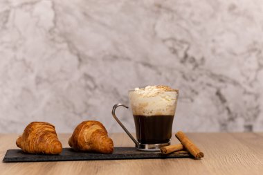Kremalı kahve, kruvasan, tarçın çubuğu ve tarçın tozu ahşap bir masada tahta bir tabakta mermer zemin, metin için boşluk