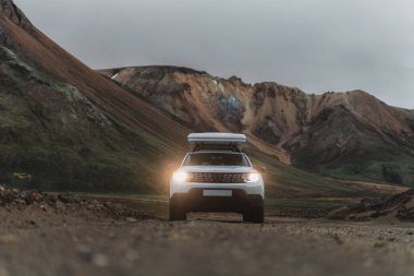 Dacia Duster 4x4 İzlanda 'da Landmannalaugar' da yol kenarında bir çatı çadırı var. Ön cephede ışıklar açık ve ön planda çakıllar var. Arka planda renkli dağları görebiliriz.